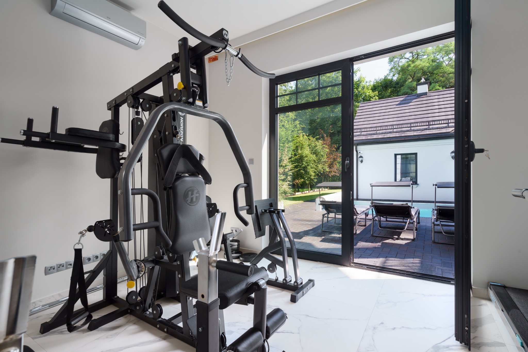 Profesjonalna siłownia domowa z wielofunkcyjnym atlasem Horizon, marmurową podłogą i widokiem na basen przez otwarte, czarne drzwi przesuwne.