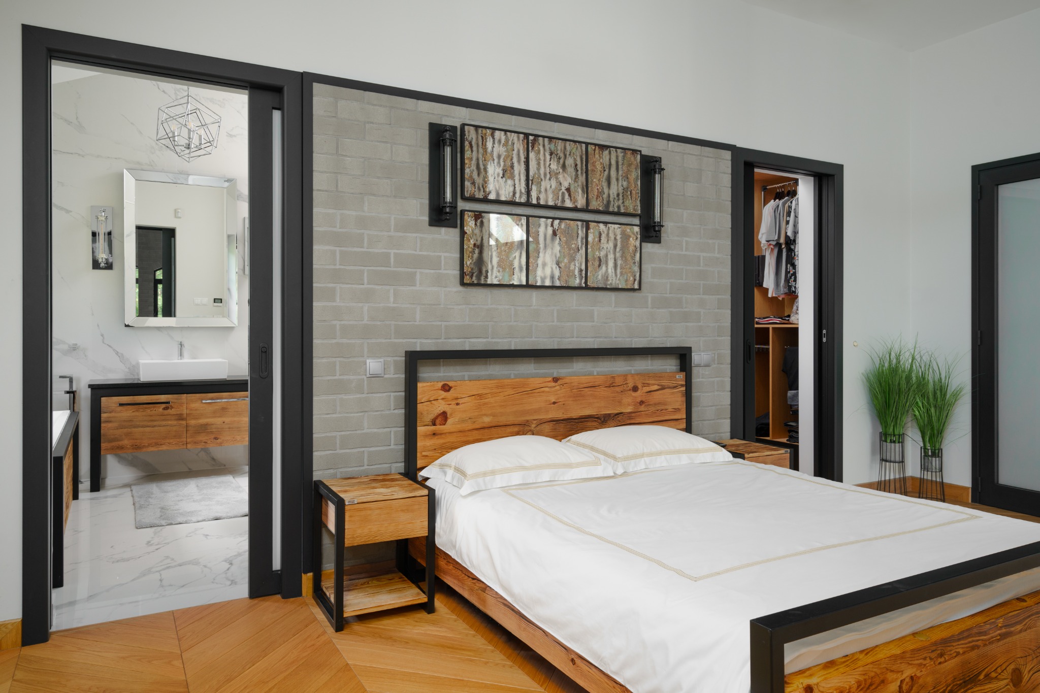 Sypialnia w stylu loft z drewnianym łóżkiem, ceglaną ścianą, widokiem na łazienkę z umywalką na szafce i garderobę z ubraniami.