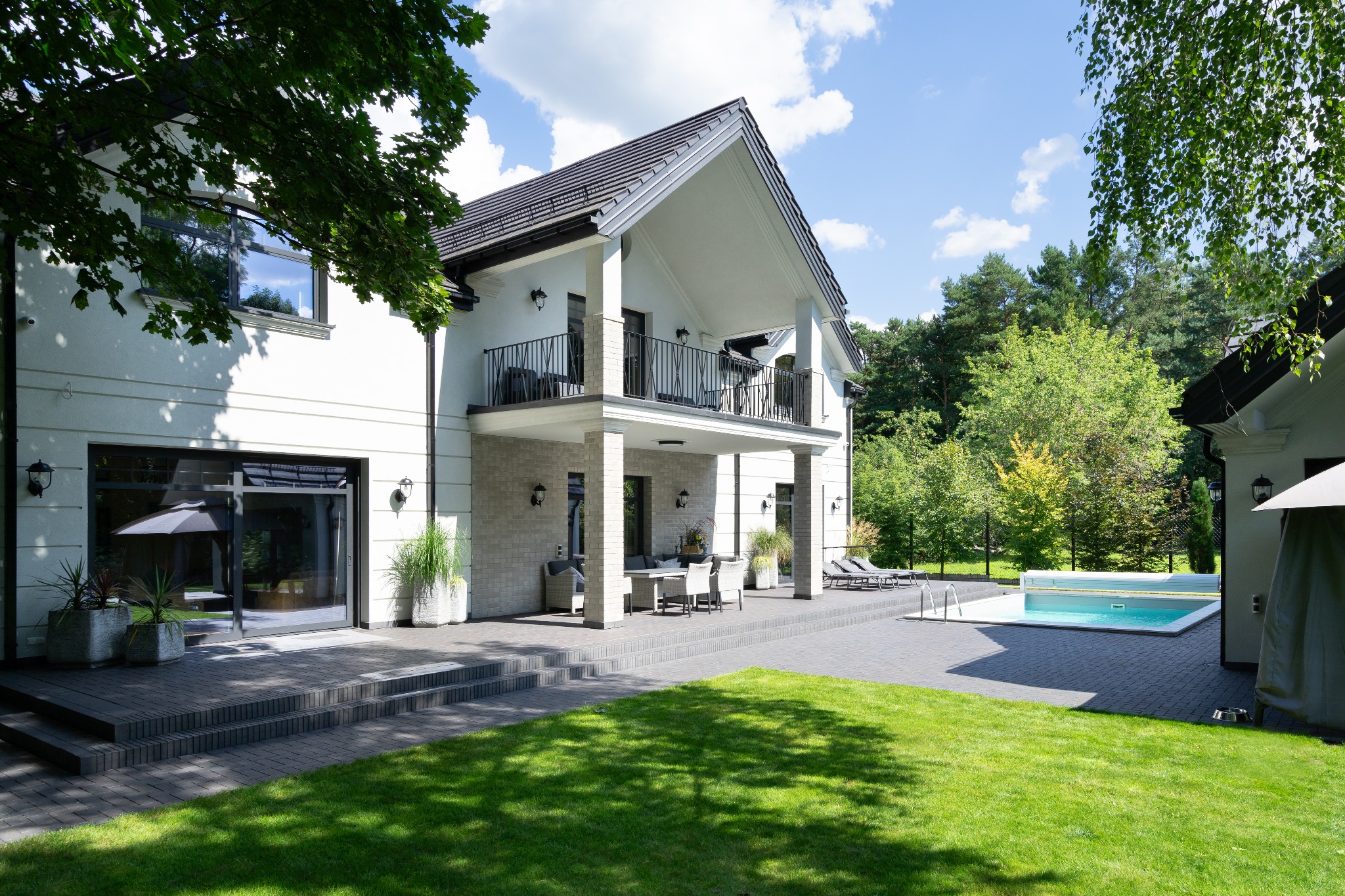 Nowoczesny dom z białymi ścianami, czarnym dachem i dużym tarasem, otoczony zielonym trawnikiem i drzewami, z widocznym basenem w tle.