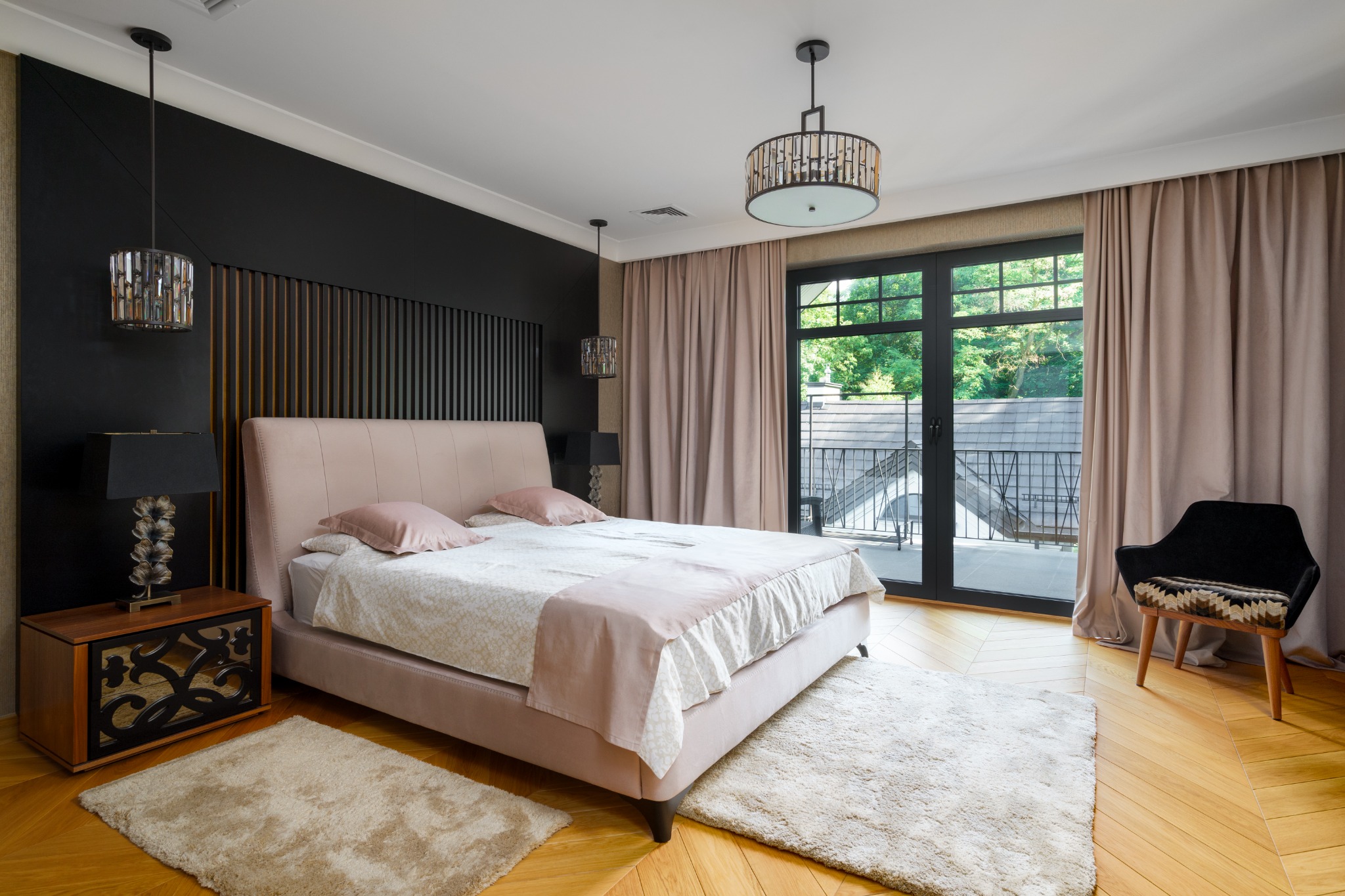 Elegancka sypialnia z łóżkiem w pudrowym różu, drewnianą podłogą w jodełkę i czarną ścianą z drewnianymi lamelami. Widok na balkon z zielenią w tle.