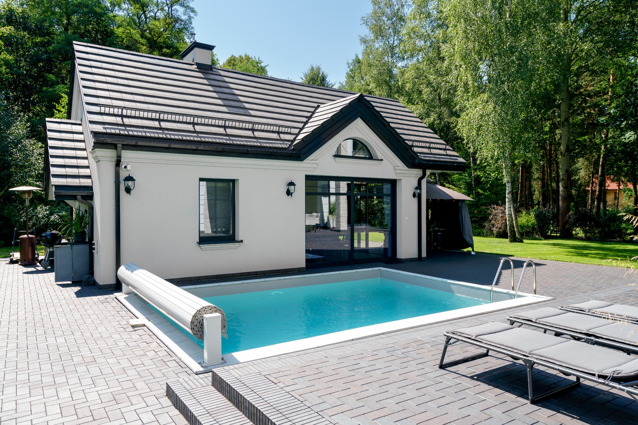 Nowoczesny dom z basenem ogrodowym o turkusowej wodzie, widoczne leżaki i zadaszenie basenu, otoczony zielenią.