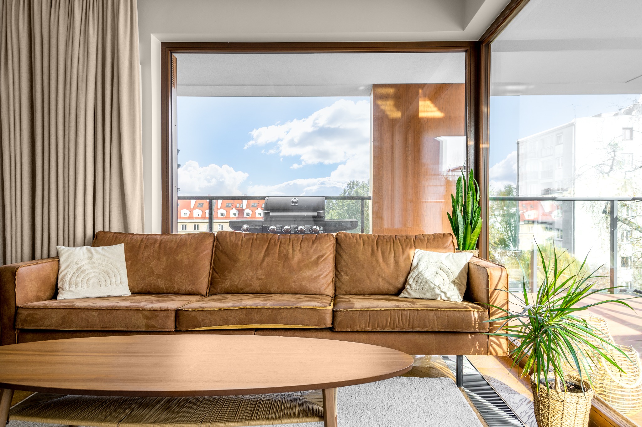 Salon z brązową skórzaną sofą, drewnianym stolikiem kawowym i widokiem na balkon z grillem gazowym.