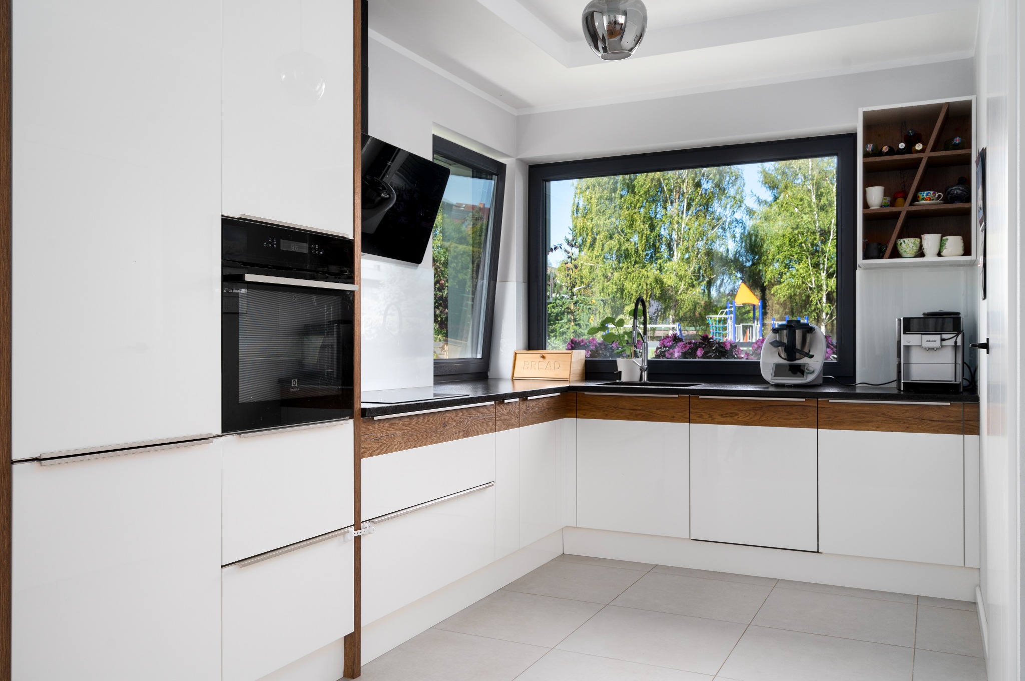 Nowoczesna kuchnia z białymi lakierowanymi frontami, czarnym blatem i drewnianymi akcentami, widok na plac zabaw przez duże okno.
