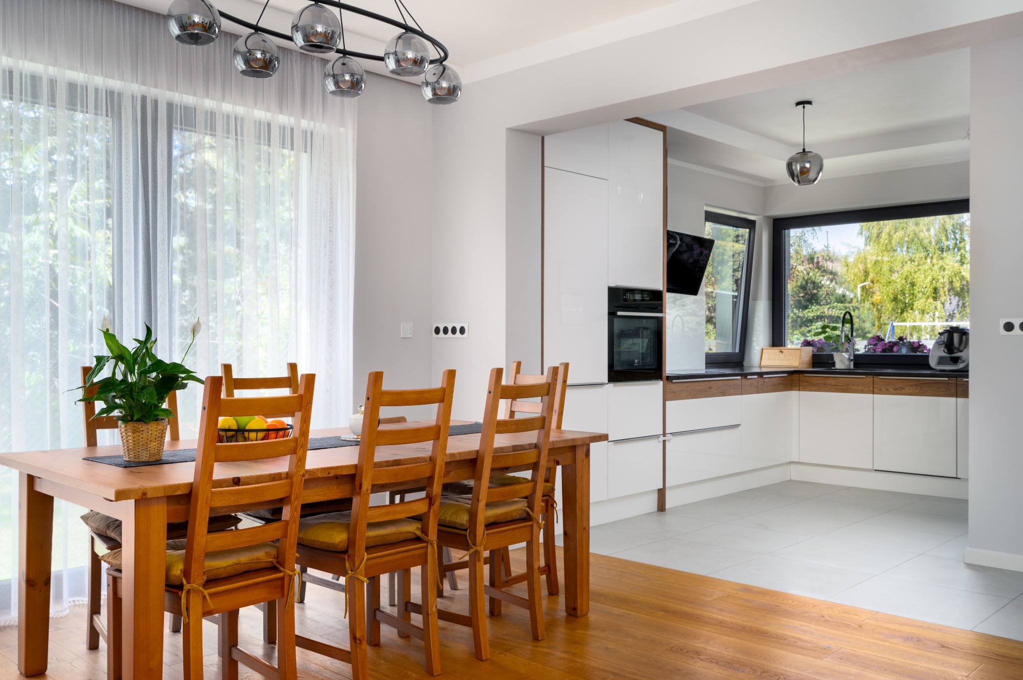 Jasna kuchnia z drewnianym stołem i krzesłami, widok na ogród przez duże okno, nowoczesne oświetlenie i minimalistyczne białe szafki kuchenne.