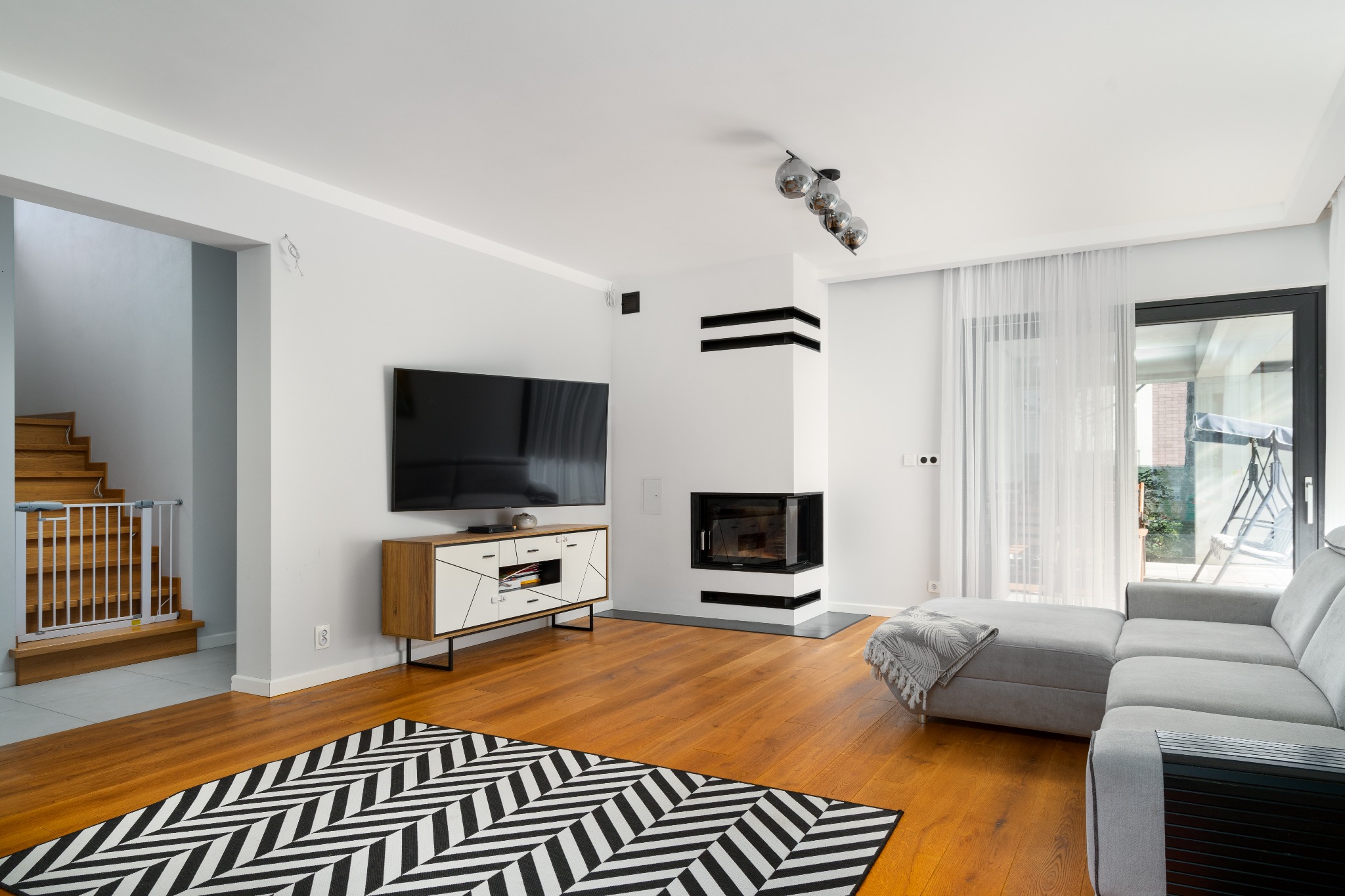 Nowoczesny salon z drewnianą podłogą, kominkiem z czarnymi akcentami, szarą sofą narożną i dywanem w zygzaki, widok na schody z barierką zabezpieczającą.