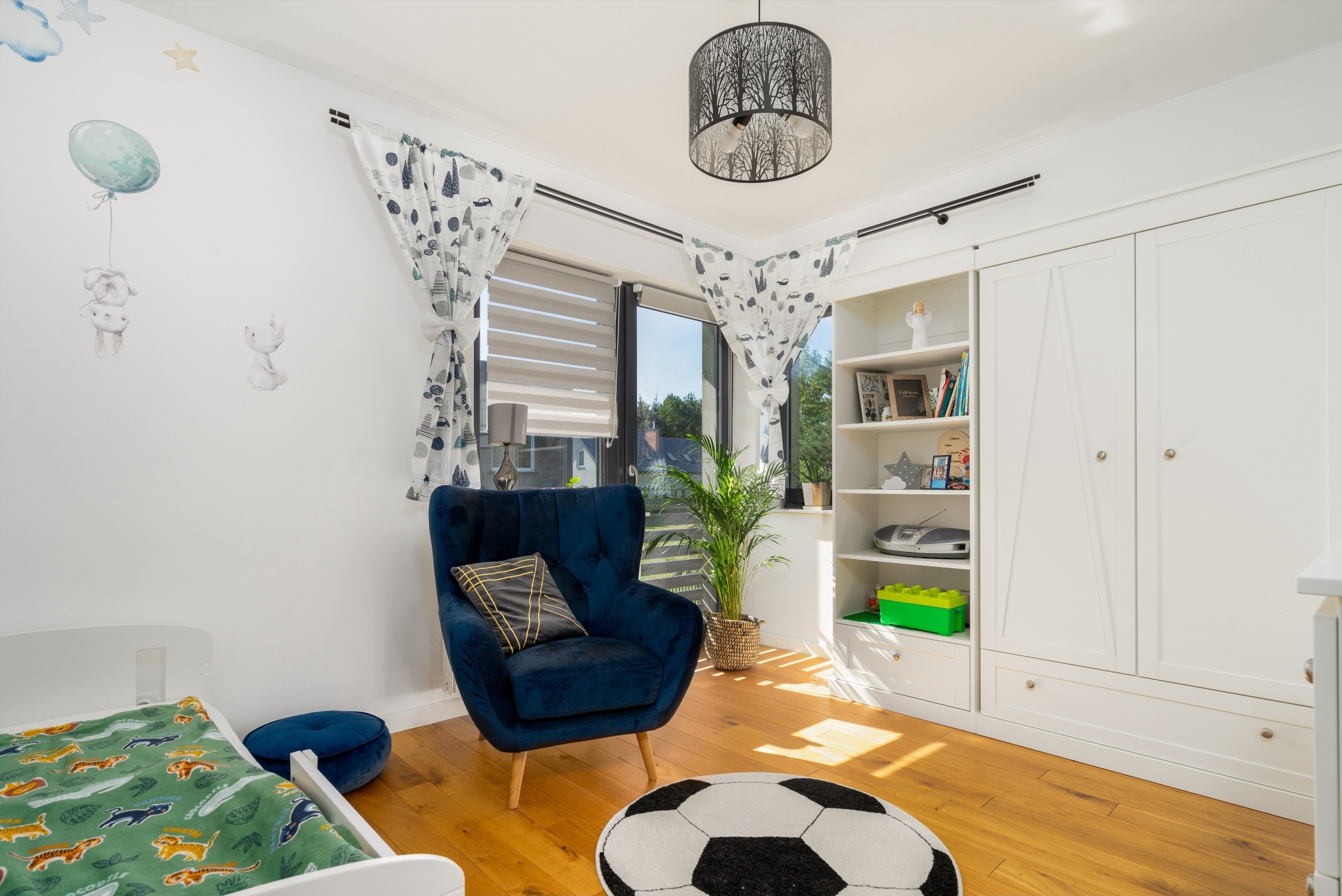 Pokój dziecięcy z białymi meblami, granatowym fotelem i dywanem w kształcie piłki nożnej, oświetlony lampą z motywem drzew.