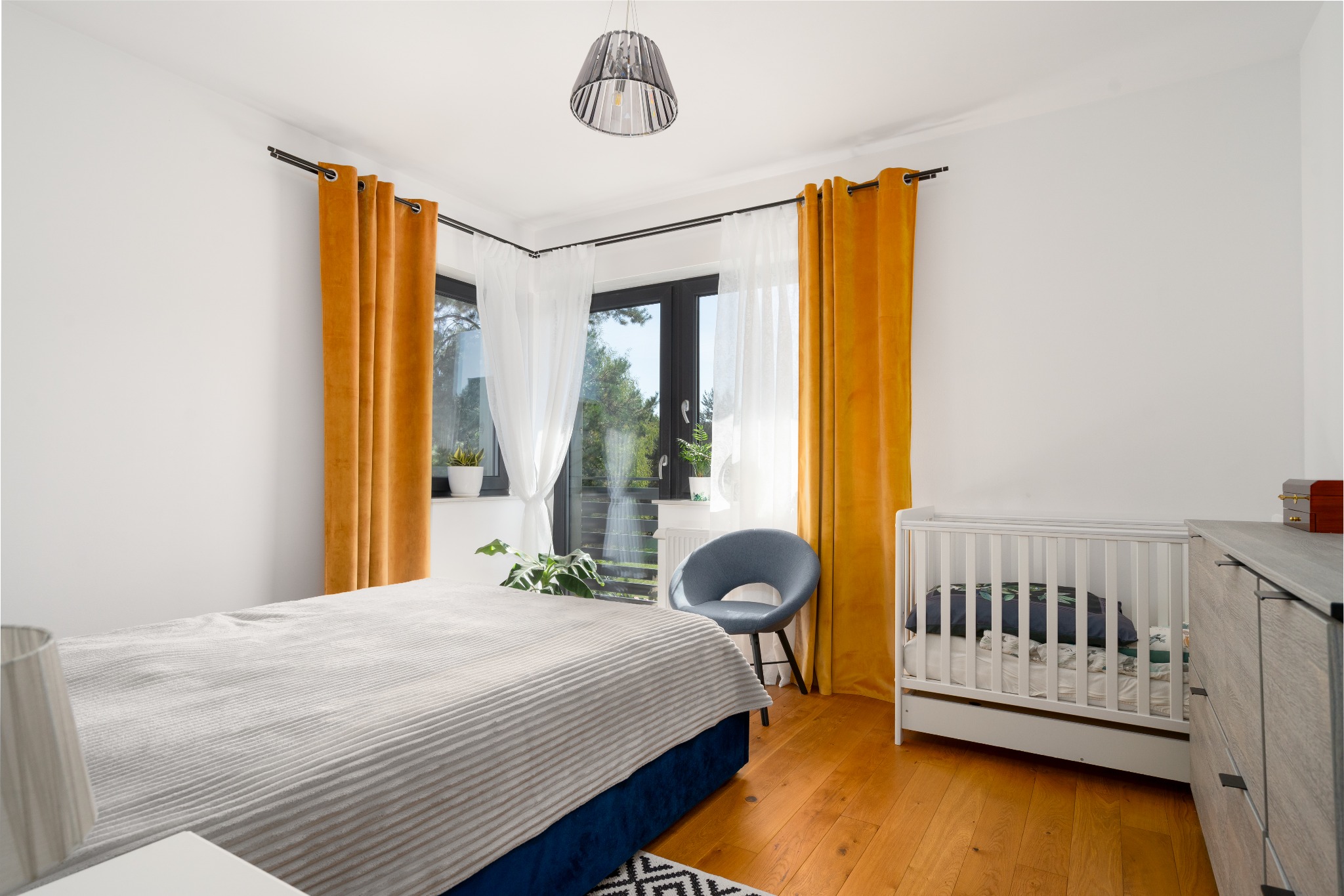 Jasna sypialnia z drewnianą podłogą, łóżkiem z niebieską ramą i szarą narzutą, białym łóżeczkiem dziecięcym, musztardowymi zasłonami, okrągłym szarym fotelem i roślinami w doniczkach na parapecie.