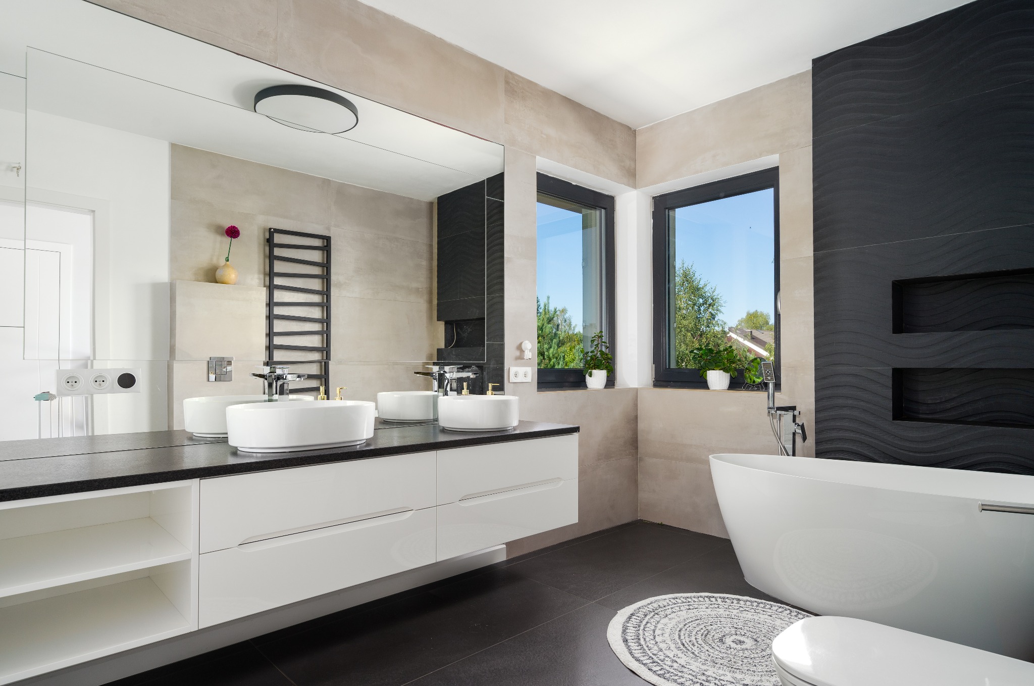 Nowoczesna łazienka z dwoma umywalkami nablatowymi, czarnym blatem, białymi szafkami, wolnostojącą wanną i czarną ścianą z dekoracyjną fakturą.
