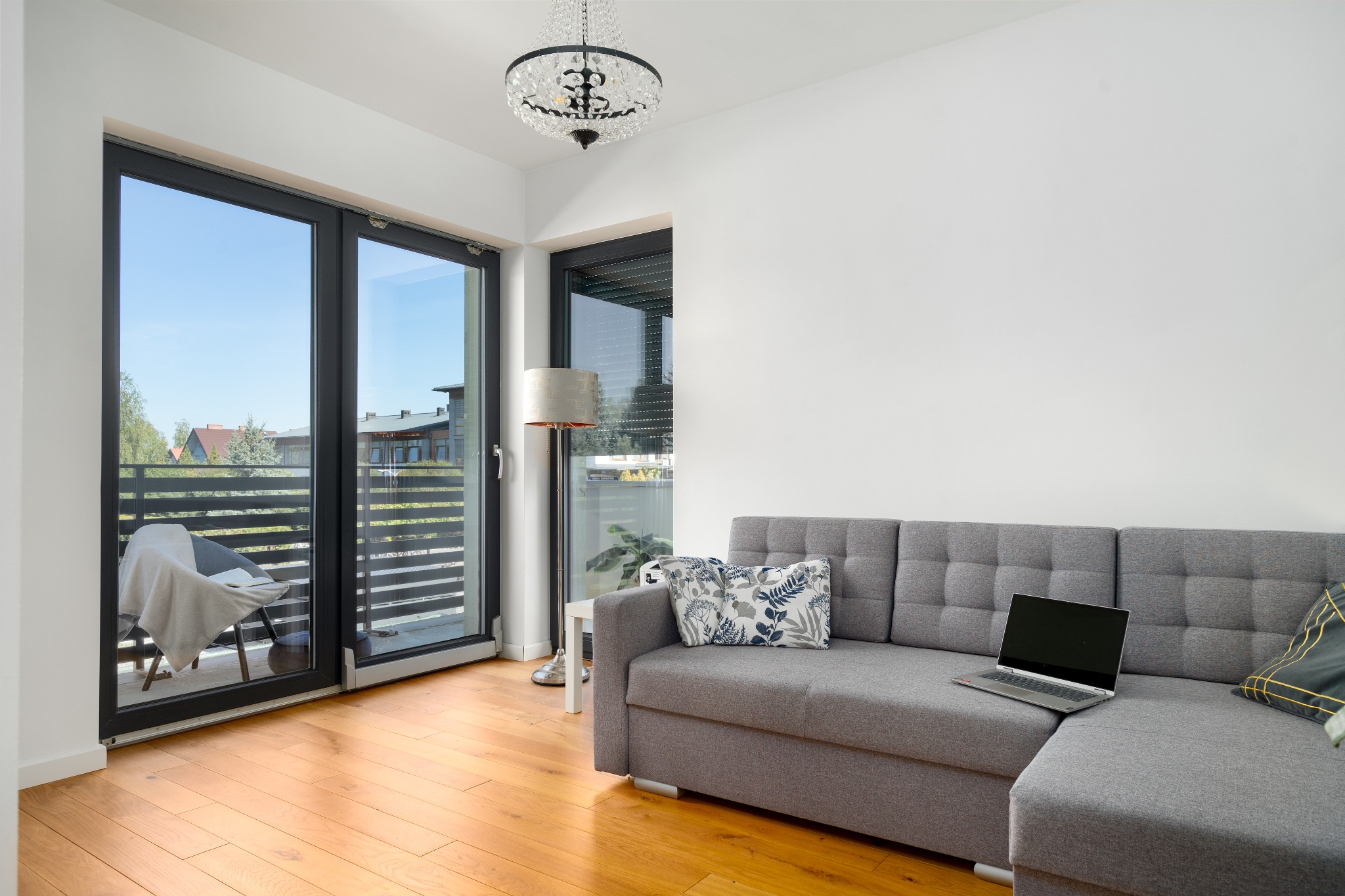 Jasne wnętrze salonu z szarą sofą narożną, otwartym laptopem, balkonem z widokiem na zieleń i kryształowym żyrandolem.