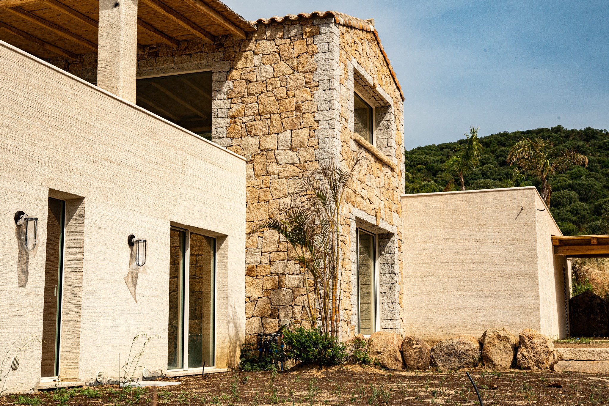 Dwukondygnacyjny dom z elewacją z jasnego kamienia i beżowego tynku o wyraźnej fakturze, z widocznymi oknami i roślinnością u podstawy. Dach kryty dachówką ceramiczną.