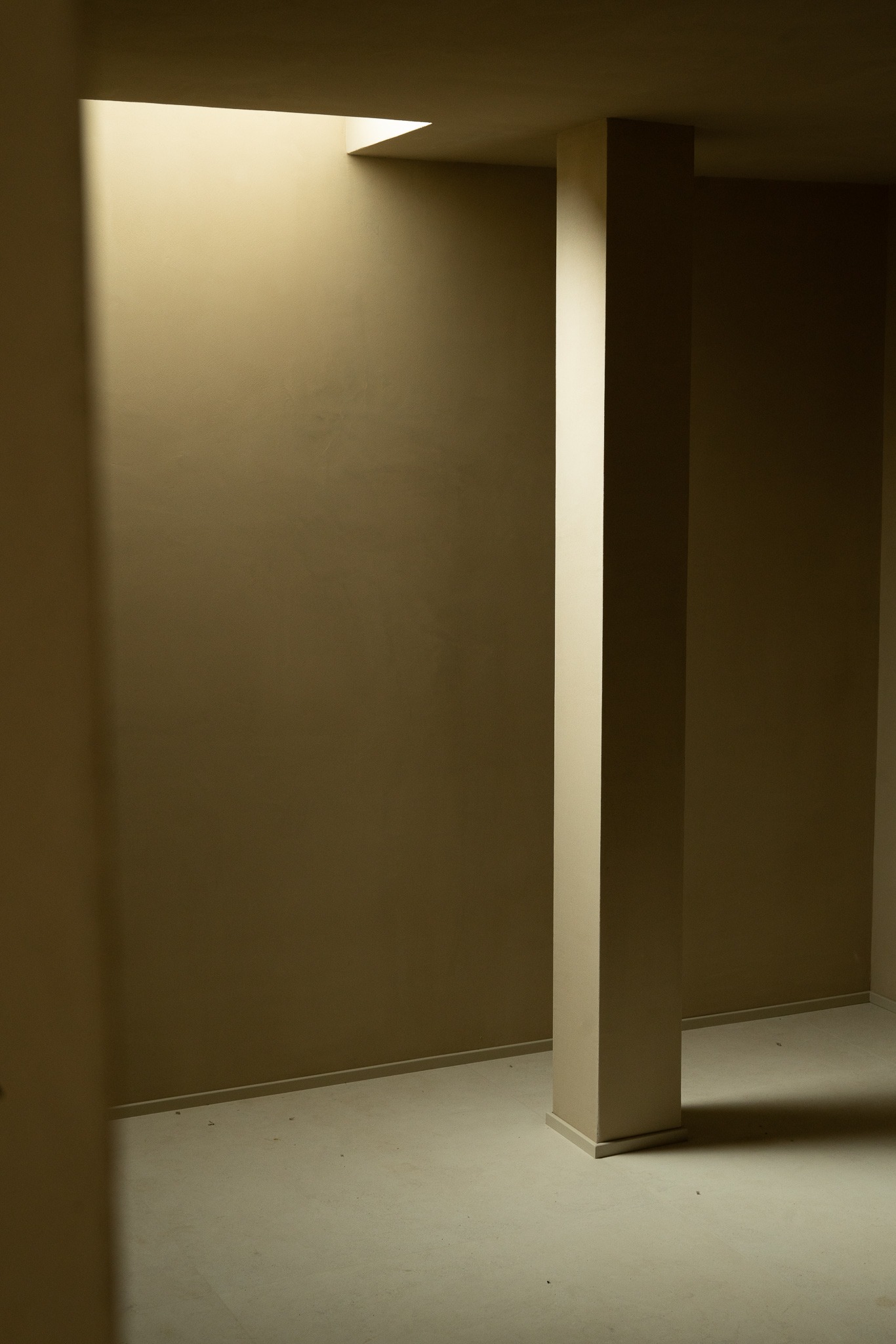 Minimalistyczne wnętrze z kolumną oświetloną przez świetlik, beżowe ściany i podłoga, gra światła i cienia.