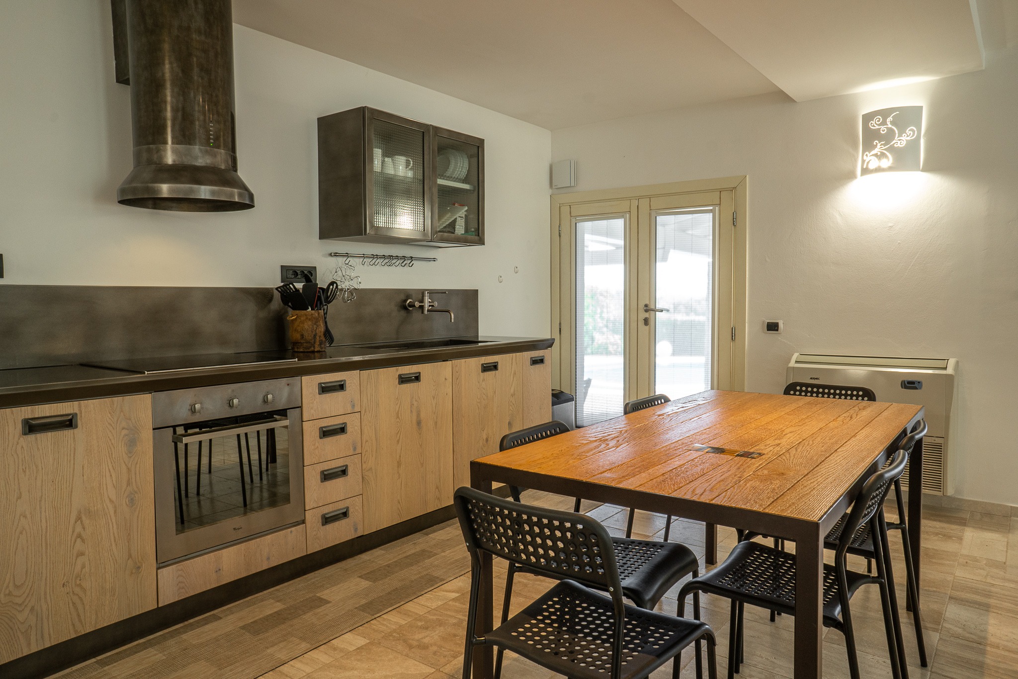 Jasna kuchnia z drewnianymi szafkami, metalowym okapem i stołem jadalnym z czarnymi krzesłami, oświetlona dekoracyjnym kinkietem.