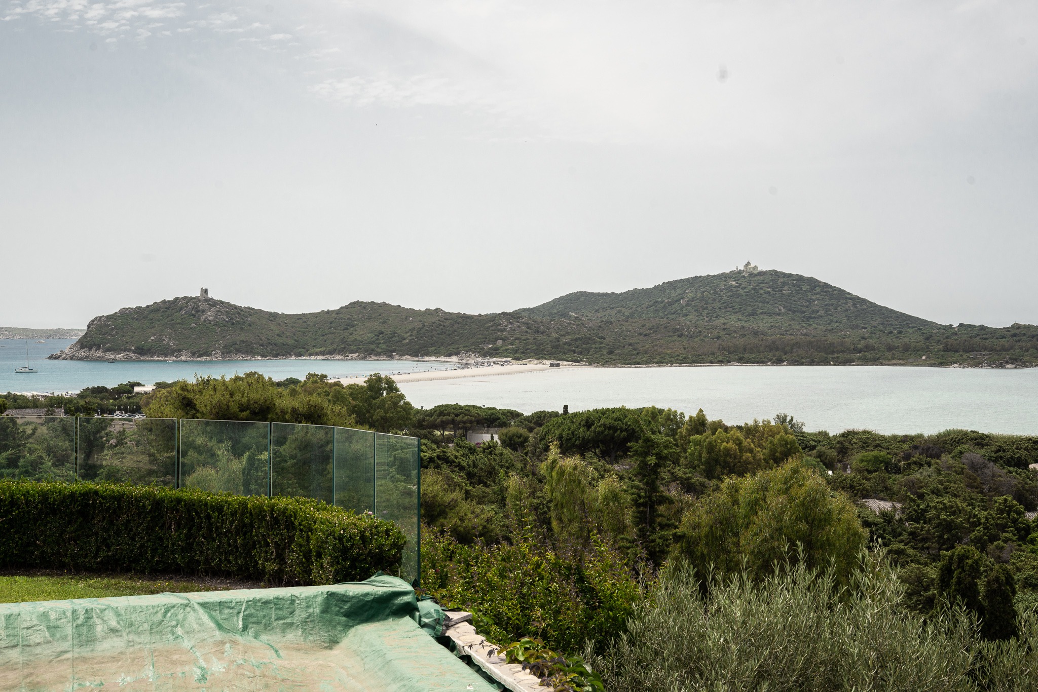 Widok z tarasu na malowniczą zatokę z piaszczystą mierzeją i zalesionymi wzgórzami zwieńczonymi wieżami, częściowo zasłonięty szklaną balustradą i zielenią.