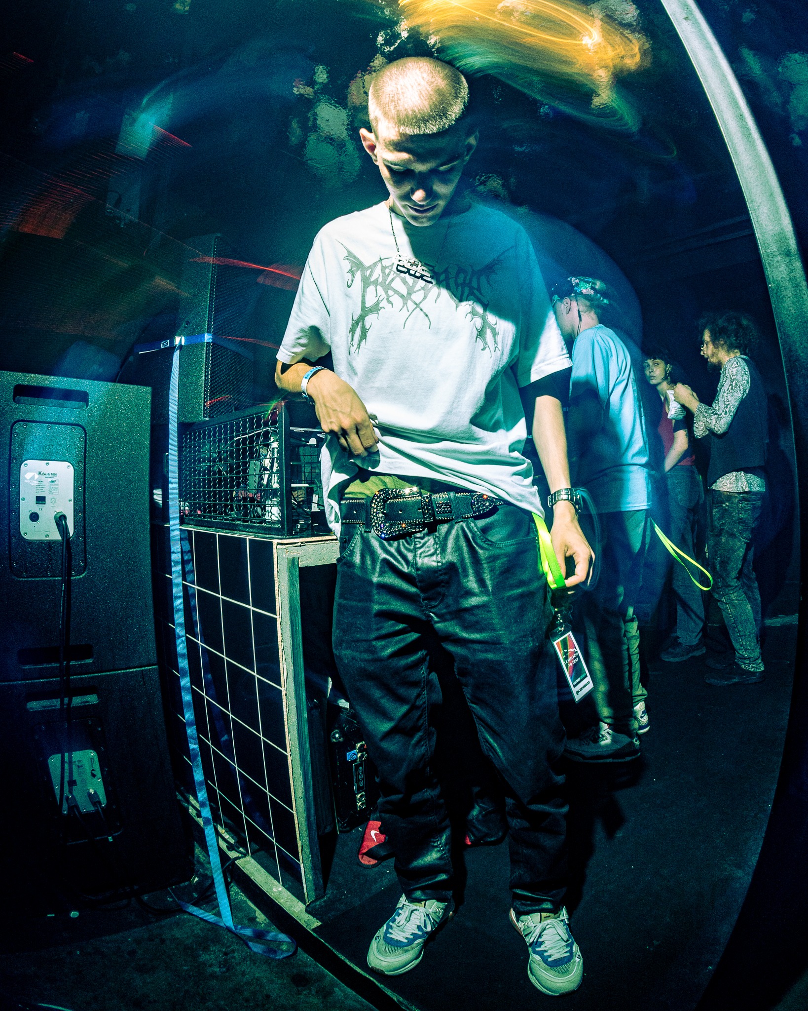 Młody mężczyzna z identyfikatorem na szyi, w koszulce z nadrukiem i ozdobnym paskiem, stoi obok kolumn głośnikowych podczas imprezy we Wrocławiu, oświetlony niebieskim światłem stroboskopowym.