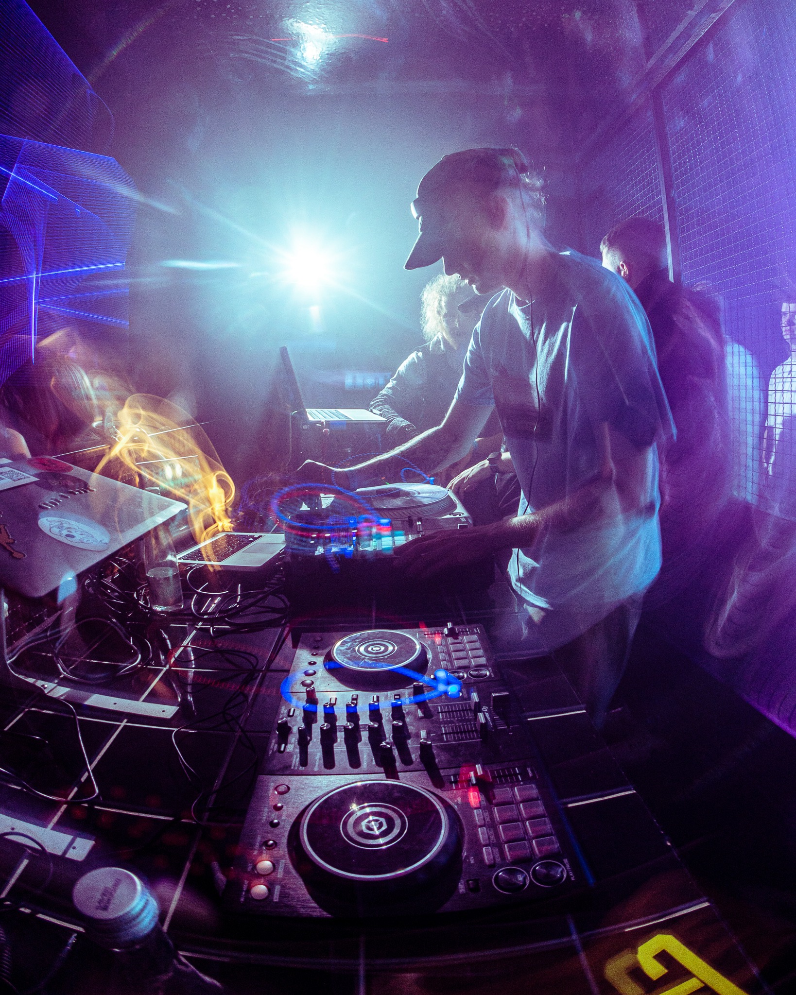 DJ miksuje muzykę na konsolecie w klubie nocnym, dynamiczne oświetlenie i efekty wizualne, widoczny laptop i butelka wody, perspektywa z dołu.