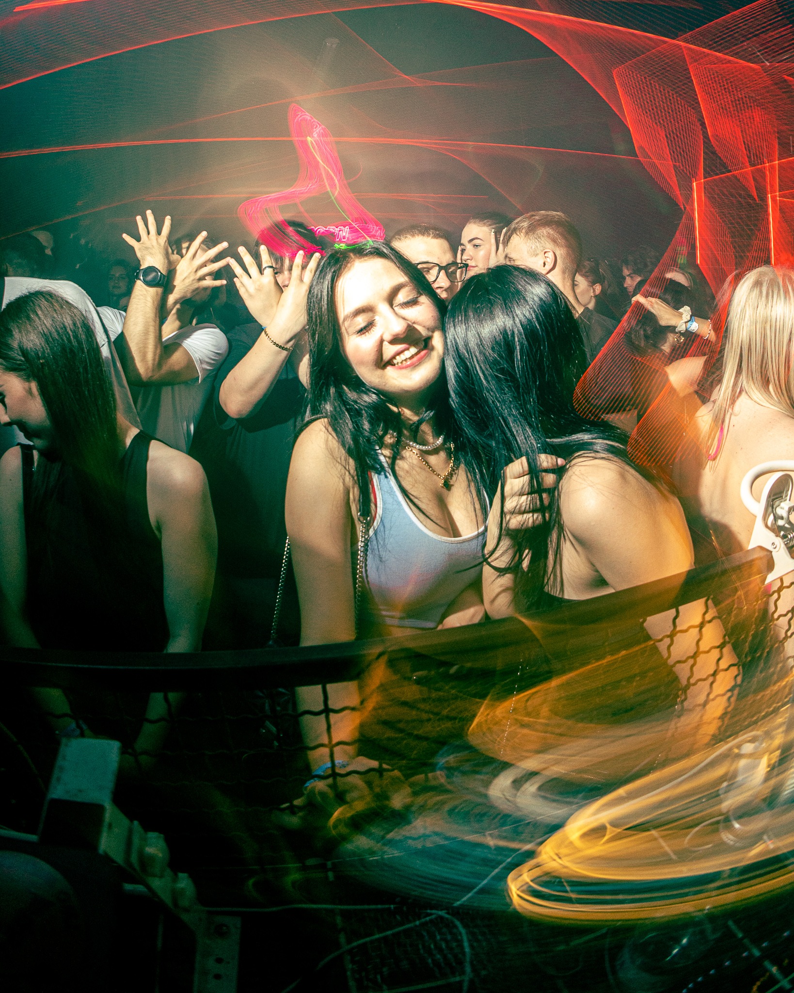 Rozmazane ujęcie z imprezy w klubie: uśmiechnięta brunetka w białym topie, złoty łańcuszek, tłum ludzi w tle, czerwone światła i ruchome smugi światła.