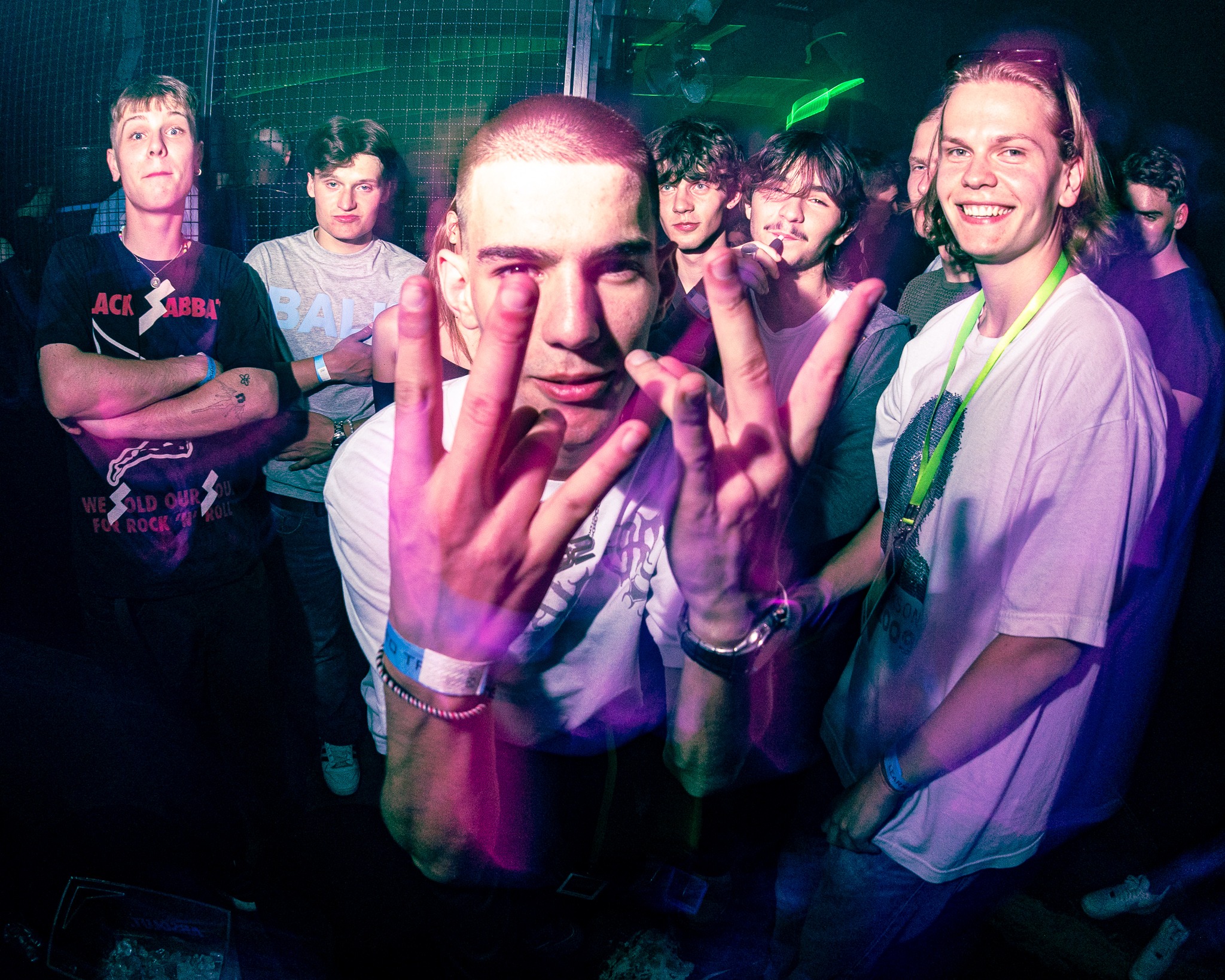 Grupa młodych osób w klubie nocnym, w tle siatka, oświetlenie neonowe, pierwszy plan mężczyzna z rękami uniesionymi w geście, widoczne opaski na rękach.