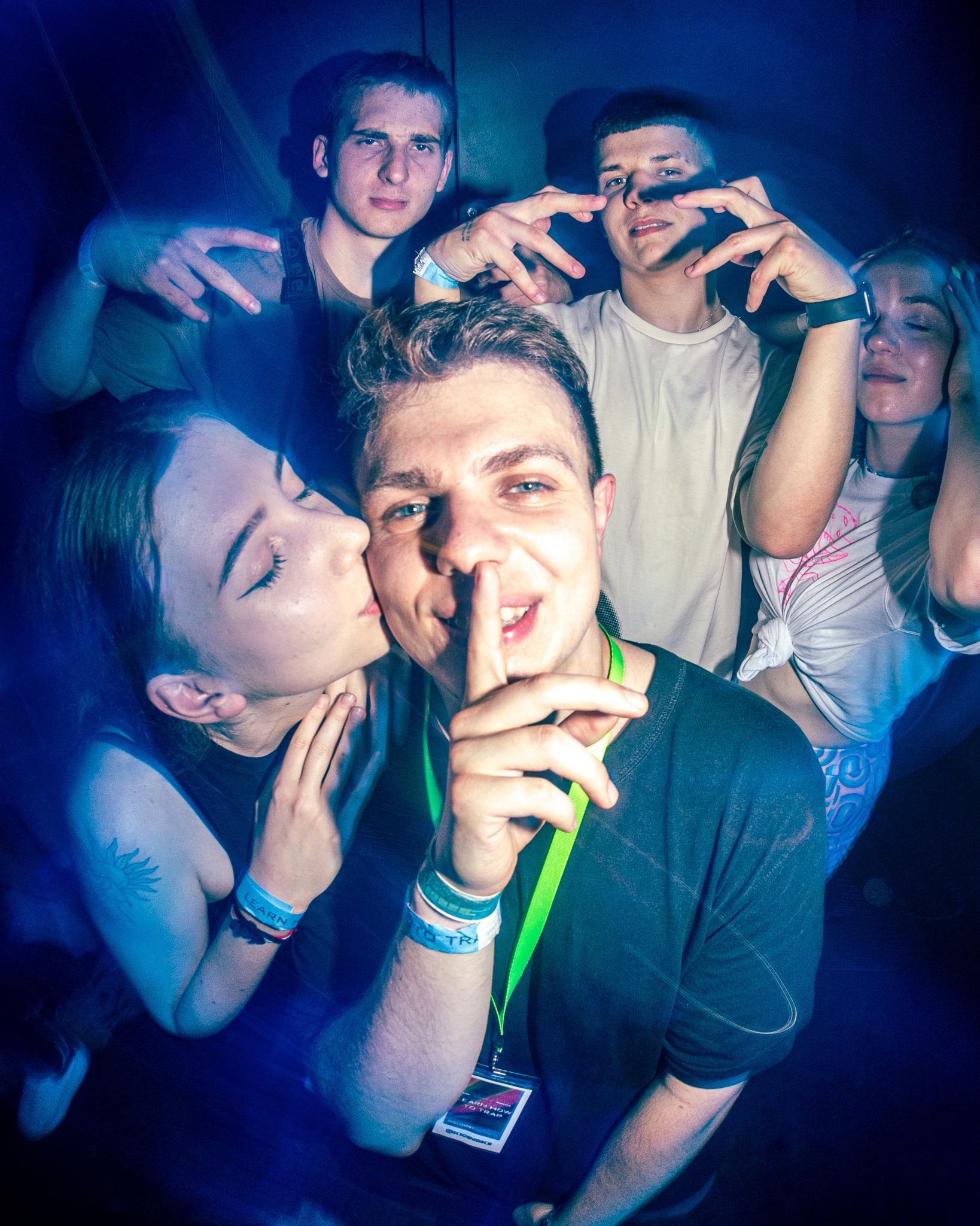 Grupa młodych ludzi pozuje do zdjęcia w niebieskim oświetleniu, dziewczyna całuje mężczyznę w policzek, a on trzyma palec na ustach w geście uciszenia; widoczne opaski na rękach, identyfikatory...