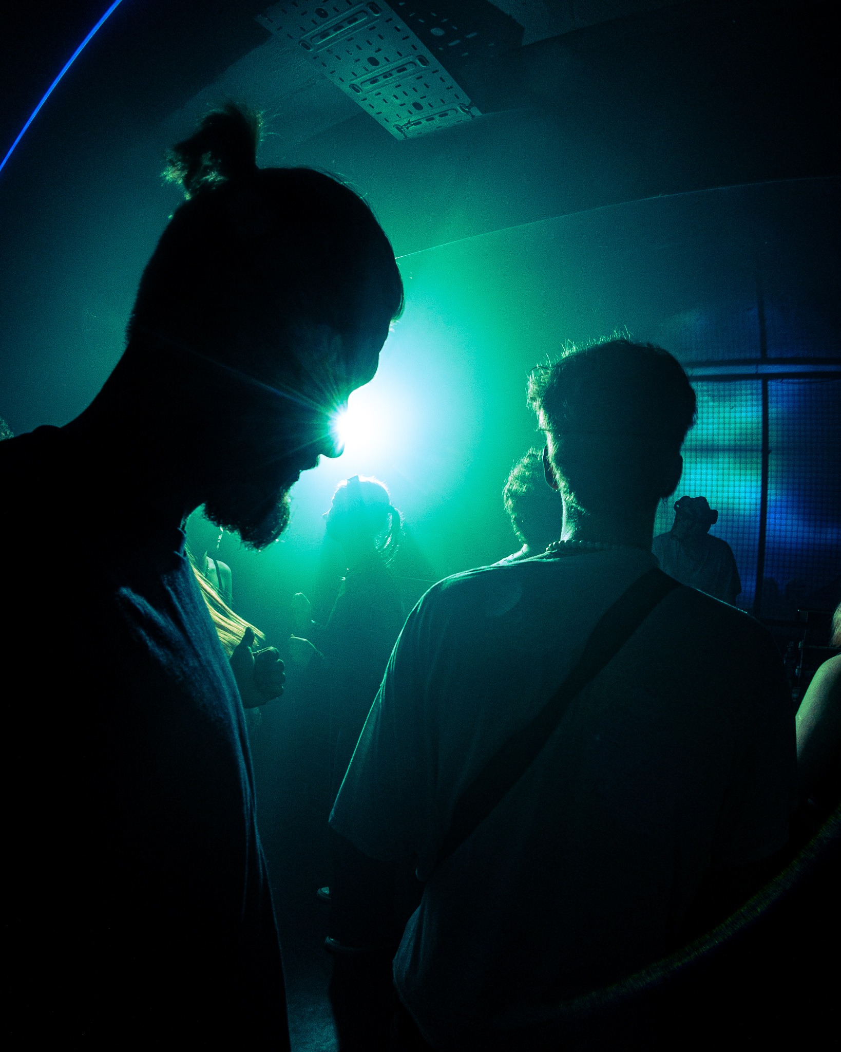 Silhouettes of people in a dark room illuminated by green and blue lights; a man with a beard and hair in a bun is in the foreground.