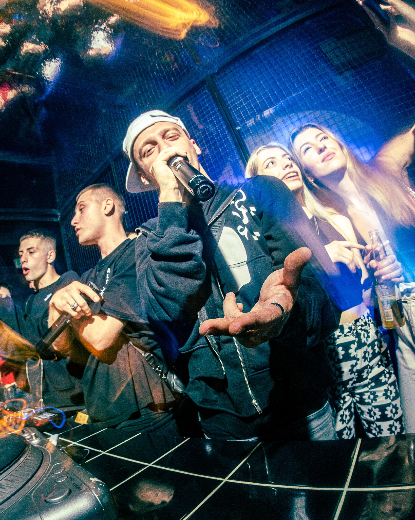 Występujący raper z mikrofonem na scenie klubu nocnego z publicznością w tle, oświetlenie sceniczne i DJ-ka widoczne.