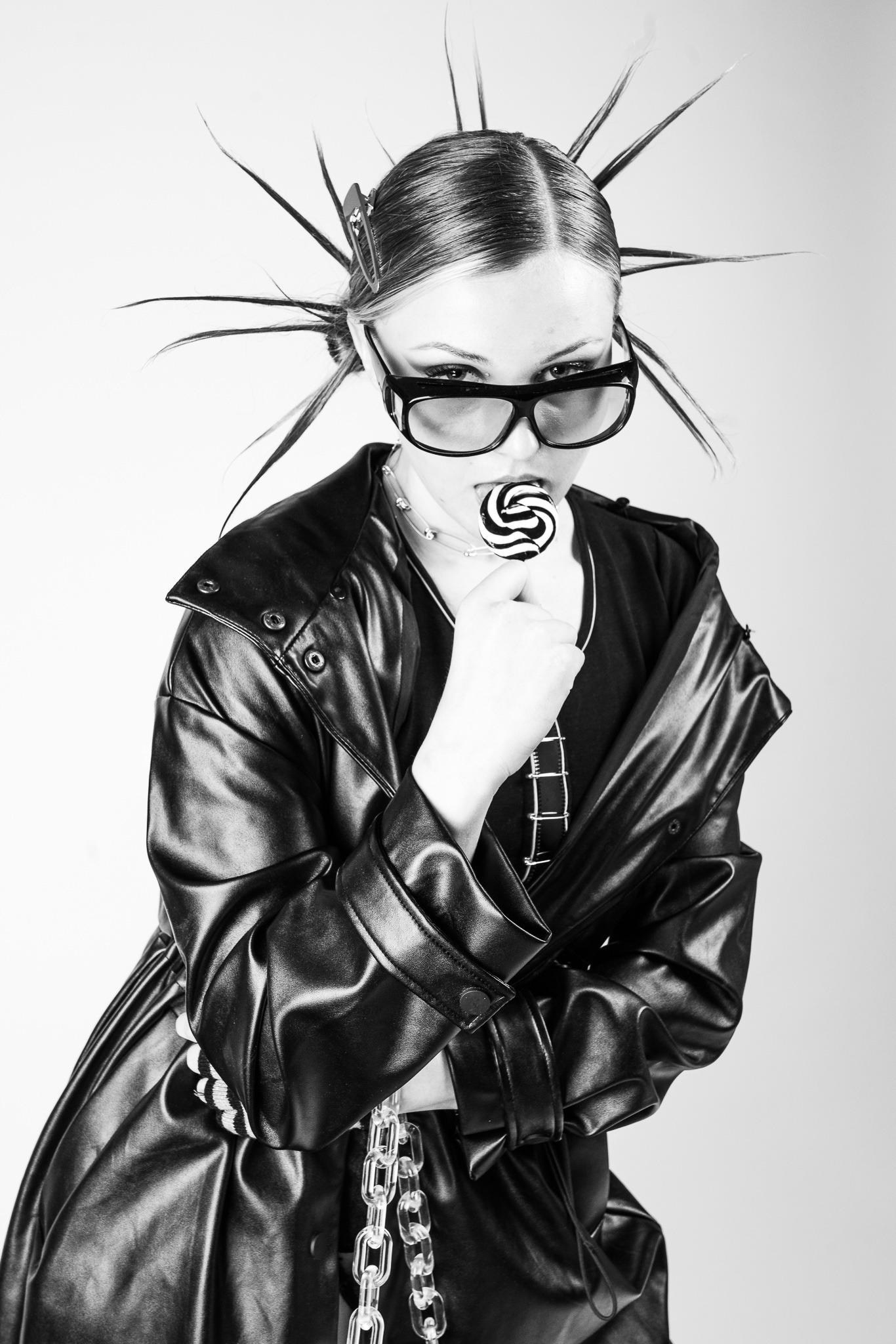 Czarno-białe portretowe ujęcie kobiety z irokezem, okularami i lizakiem w ustach, ubrana w błyszczący, skórzany płaszcz z łańcuchem.
