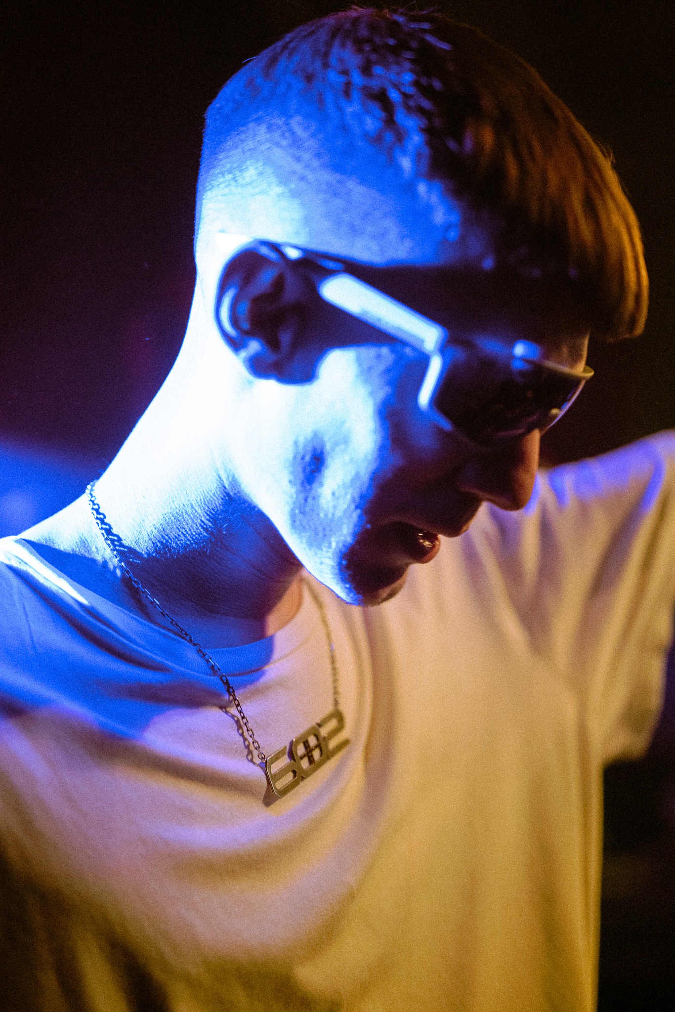 Portret młodego mężczyzny w okularach, oświetlonego niebieskim światłem, z wisiorkiem na szyi, w białej koszulce, z krótką fryzurą.