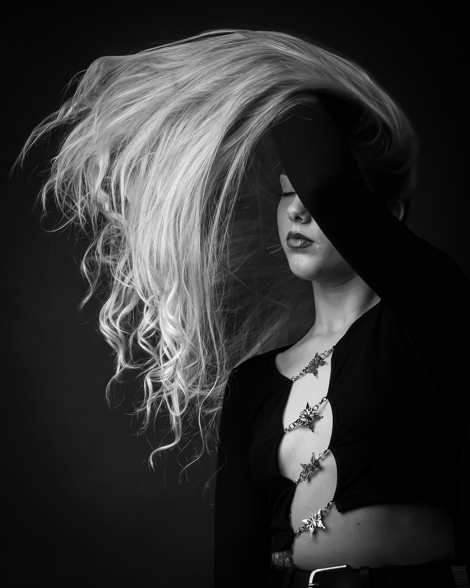 Czarno-białe zdjęcie kobiety z długimi, blond włosami zakrywającymi część twarzy, ubrana w czarną bluzkę z wycięciami zdobioną łańcuszkiem z motylami.