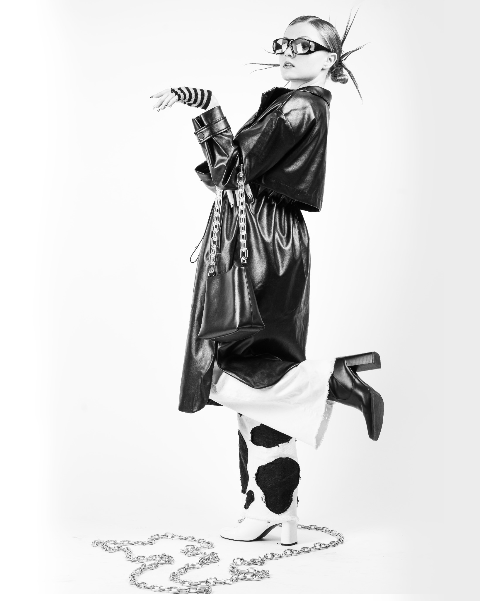 Czarno-białe zdjęcie modelki w stylizacji z czarną skórzaną kurtką, spodniami w krowie łaty, butami na obcasie i okularami, pozującej na jednej nodze z łańcuchem na podłodze.