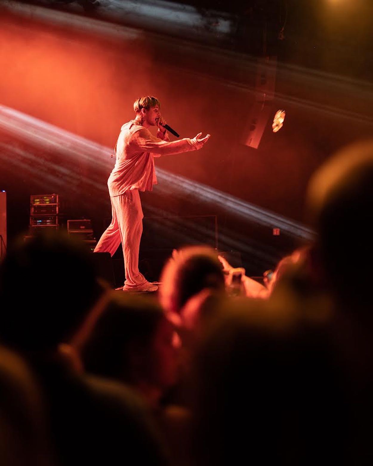 Piosenkarz w jasnym garniturze na scenie, trzymający mikrofon i gestykulujący do publiczności w czerwonym oświetleniu, z widocznymi głośnikami i częścią widowni.