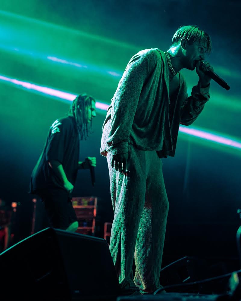 Wokalista w dzianinowym komplecie na scenie, oświetlony zielonym światłem, z drugim muzykiem w tle.