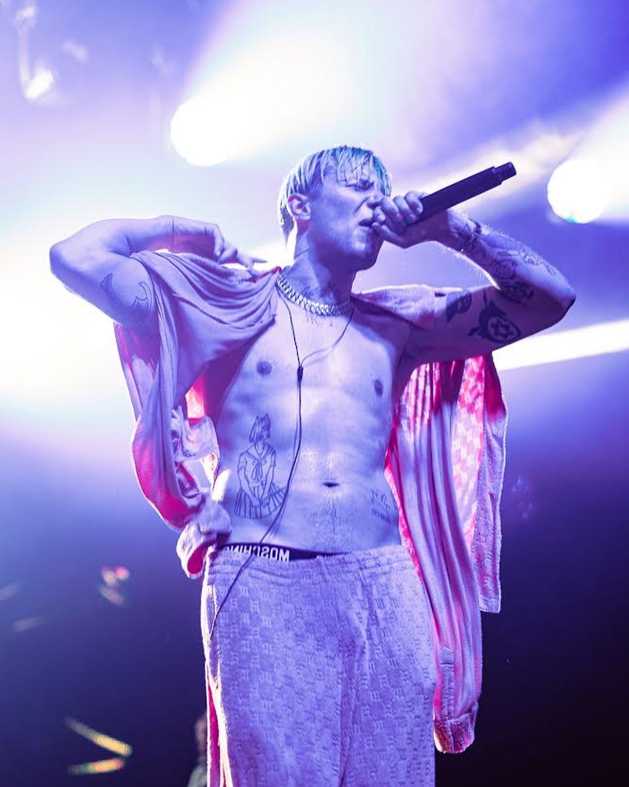 Mężczyzna z tatuażami śpiewa do mikrofonu na scenie w fioletowym oświetleniu, ubrany w różowe spodnie i narzutkę, widoczny fragment bielizny marki Moschino.