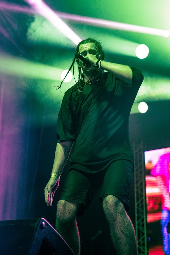 Młody raper z warkoczykami na scenie, trzyma mikrofon w dłoni, oświetlony zielonym i fioletowym światłem.