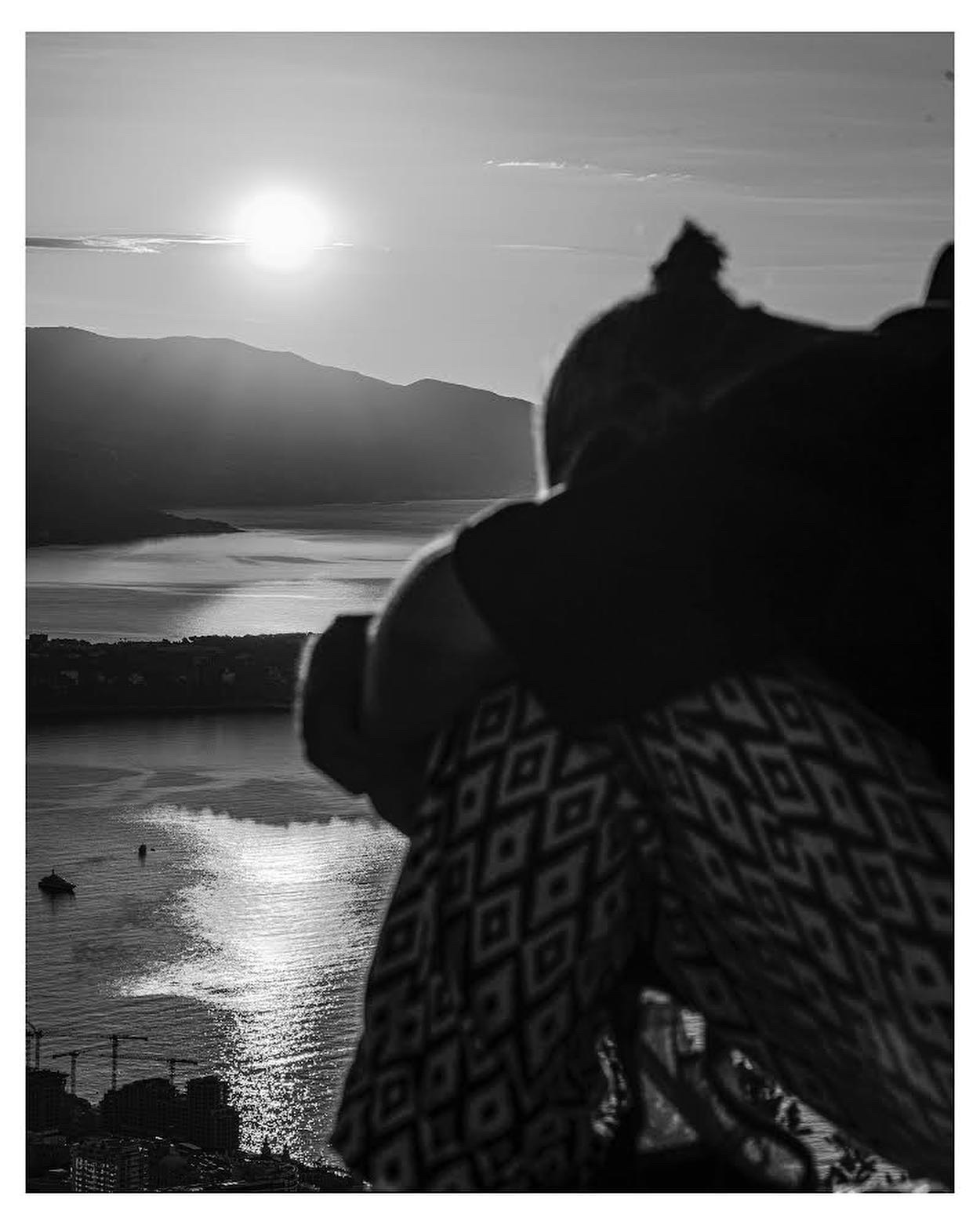 Czarno-białe zdjęcie osoby siedzącej z podciągniętymi kolanami, z widokiem na jezioro i góry w tle, oświetlone zachodzącym słońcem, z widocznymi żurawiami budowlanymi w oddali.