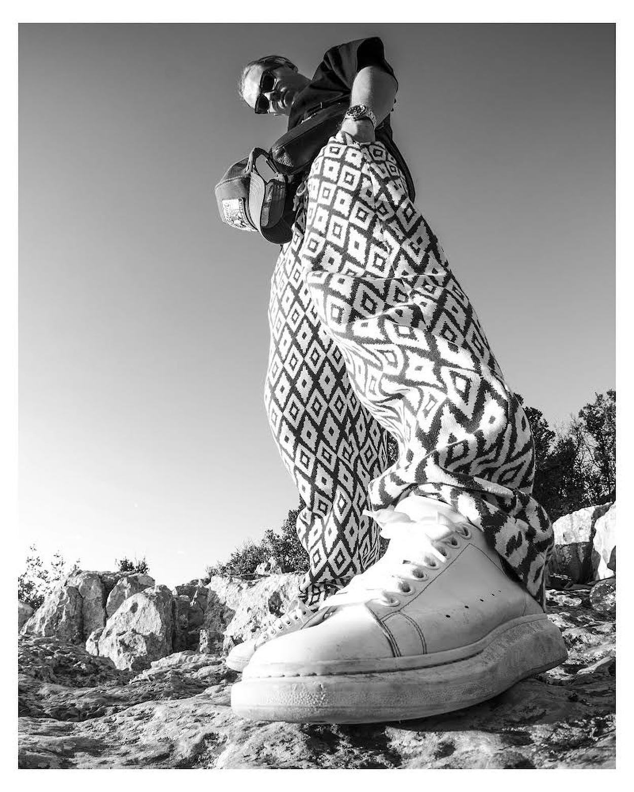 Czarno-białe zdjęcie osoby z dołu, ubrana w wzorzyste spodnie i białe buty, stojąca na skalistym terenie z torbą przewieszoną przez ramię, skierowana w dół.