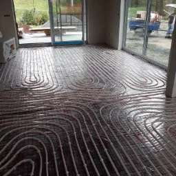 Rozłożone rury ogrzewania podłogowego na folii termoizolacyjnej w nowym budynku, widoczne okna tarasowe i materiały budowlane na zewnątrz.