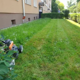 Częściowo skoszony trawnik przed blokiem mieszkalnym, z widoczną kosiarką w lewym dolnym rogu i mężczyzną przycinającym żywopłot w tle.