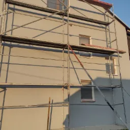 Świeżo otynkowana elewacja domu jednorodzinnego w kolorze szaro-beżowym, z oknami zabezpieczonymi folią i rozstawionym rusztowaniem, widziana w popołudniowym słońcu.