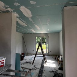 Oskar Pykało - Wnętrze w trakcie remontu: sufit z płyt gipsowo-kartonowych z widocznymi łączeniami pokrytymi szpachlą, drabina, przewody elektryczne na podłodze, rusztowanie w rogu pokoju, okno z widokiem na zieleń.