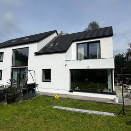 Oskar Pykało - Nowoczesny dom z białymi ścianami i czarnymi ramami okiennymi w Białymstoku, widok z ogrodu, widoczne ślady po pracach wykończeniowych elewacji oraz narzędzia.