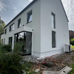 Nowoczesny, dwukondygnacyjny dom z białymi ścianami i czarnymi ramami okiennymi; widoczne ślady prac wykończeniowych na gruncie.