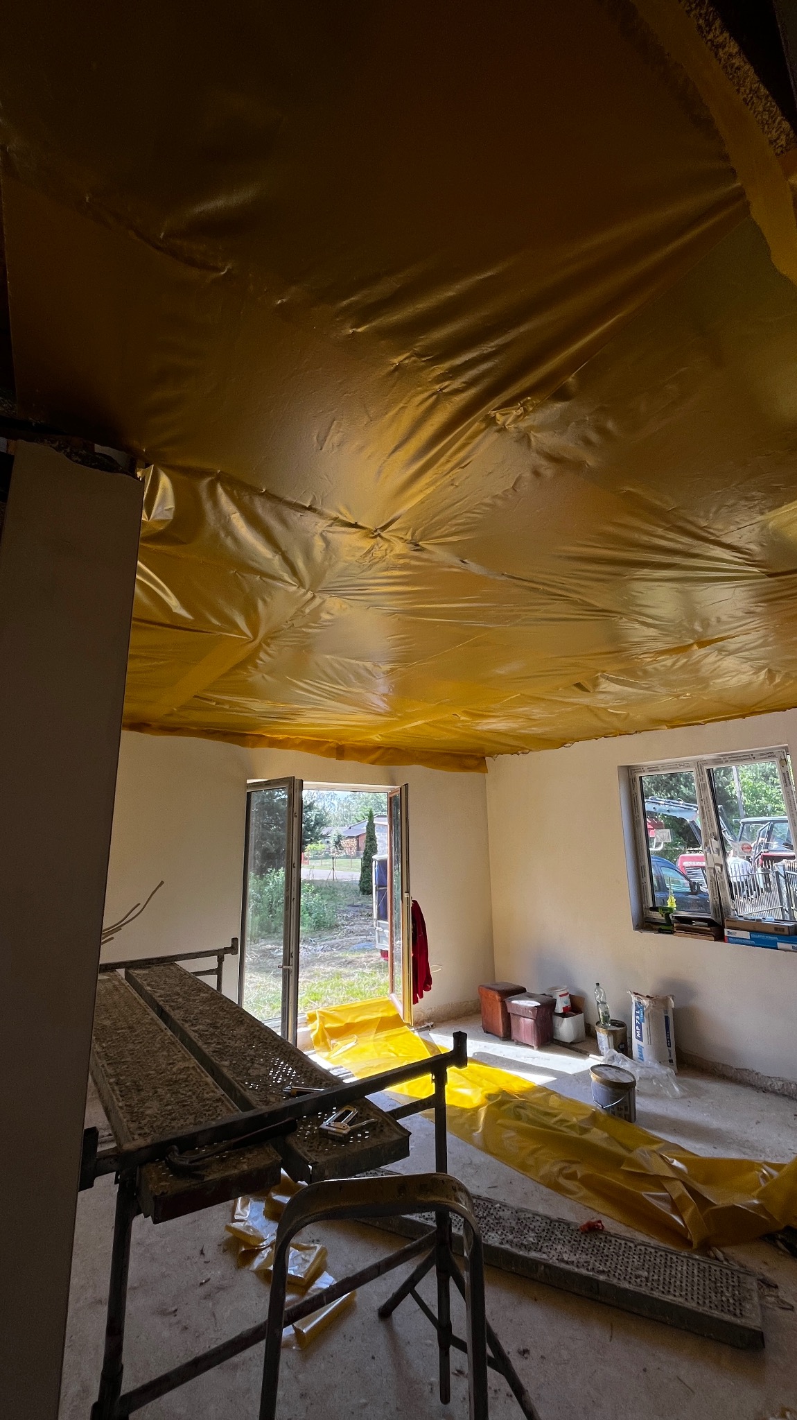 Wnętrze pokoju zabezpieczone folią malarską, widoczne rusztowanie, okno i drzwi balkonowe.