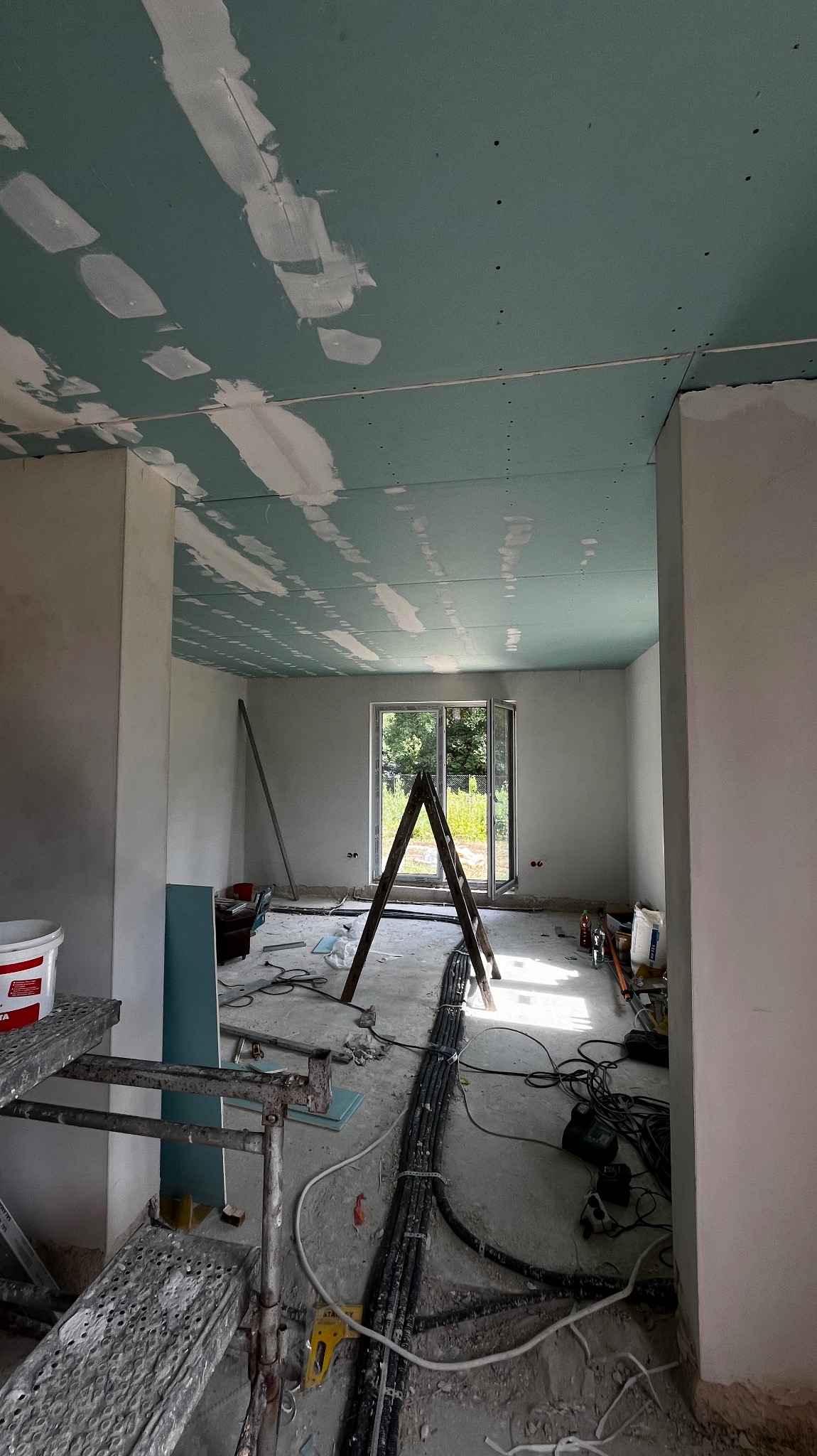 Wnętrze w trakcie remontu: sufit z płyt gipsowo-kartonowych z widocznymi łączeniami pokrytymi szpachlą, drabina, przewody elektryczne na podłodze, rusztowanie w rogu pokoju, okno z widokiem na zieleń.