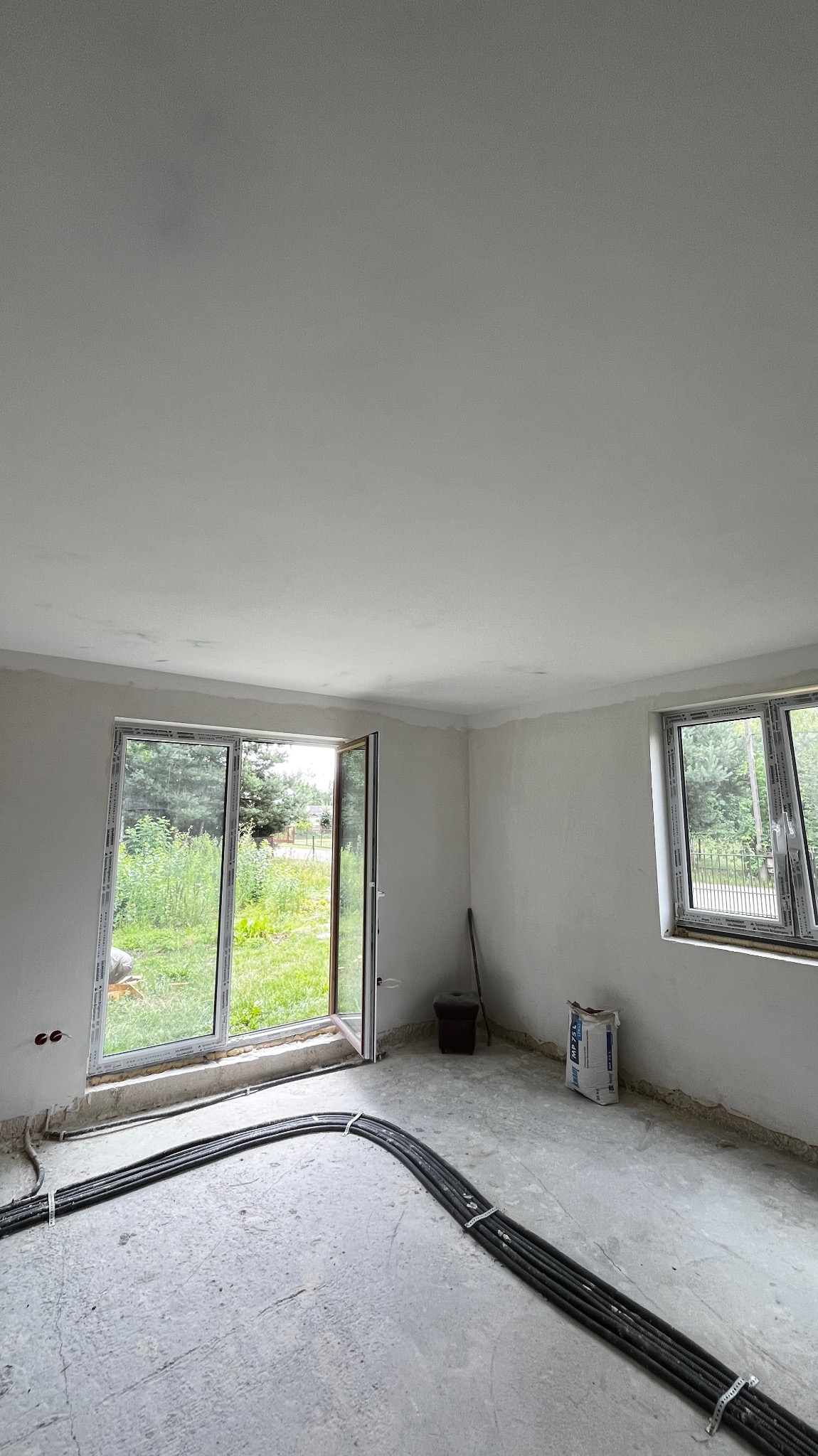 Wnętrze w trakcie remontu: surowa podłoga, świeżo zagruntowane ściany, otwarte drzwi balkonowe z widokiem na zieleń, wiązka kabli elektrycznych na podłodze, worek gładzi MP 75L przy ścianie.