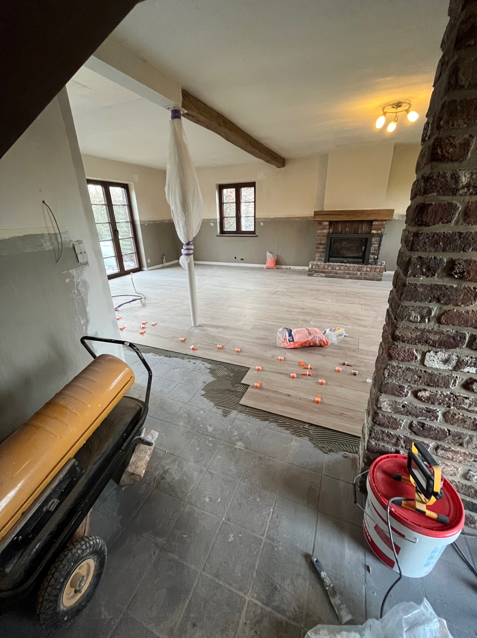 Układanie płytek podłogowych w przestronnym salonie z kominkiem, widoczny nagrzewnica i wiadro z narzędziami, płytki w trakcie montażu z użyciem klipsów poziomujących.