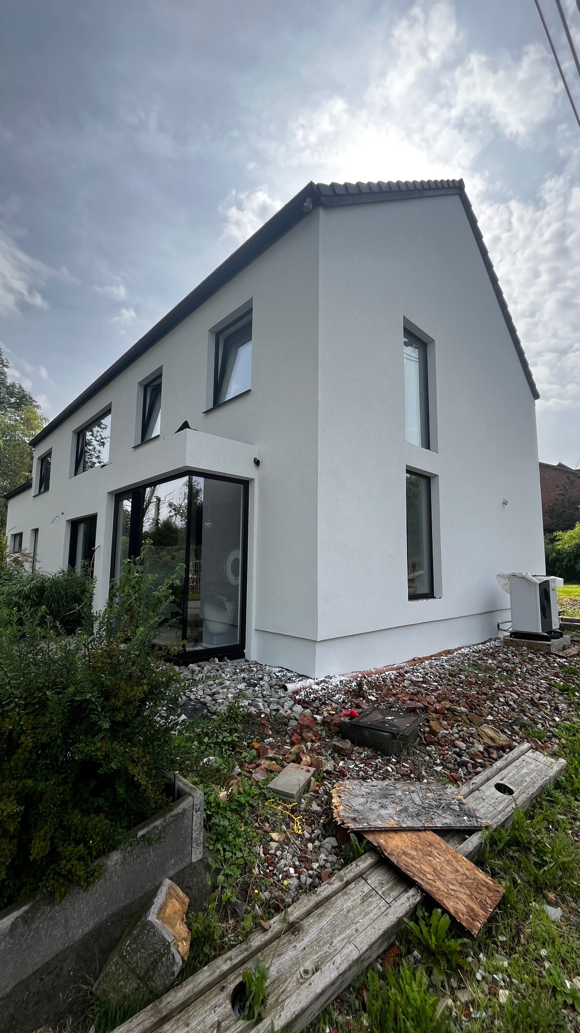 Nowoczesny, dwukondygnacyjny dom z białymi ścianami i czarnymi ramami okiennymi; widoczne ślady prac wykończeniowych na gruncie.