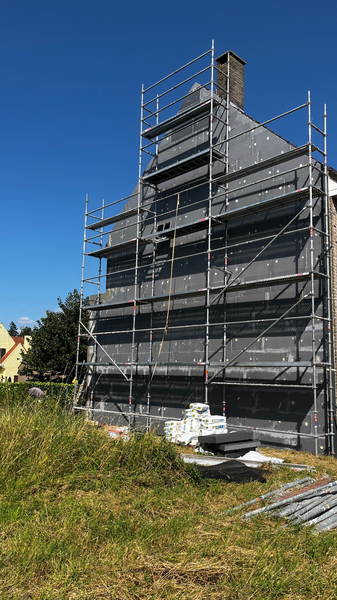 Dom w trakcie prac izolacyjnych: szare płyty styropianowe z białymi kołkami zamocowane na elewacji, rusztowanie obejmujące całą ścianę, worki z materiałem budowlanym u podstawy ściany, wysoka trawa...