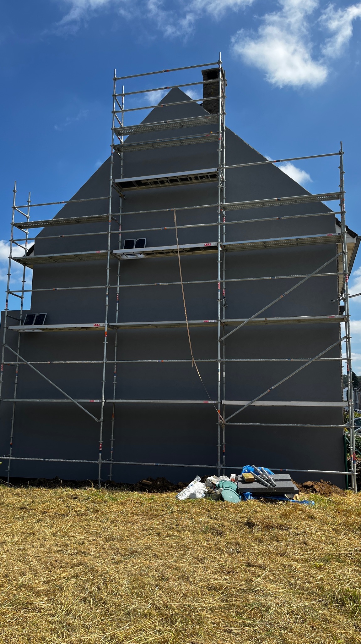 Dom w trakcie prac elewacyjnych, otoczony rusztowaniem, z widocznym fragmentem niezagospodarowanego terenu i materiałami budowlanymi u podnóża ściany.