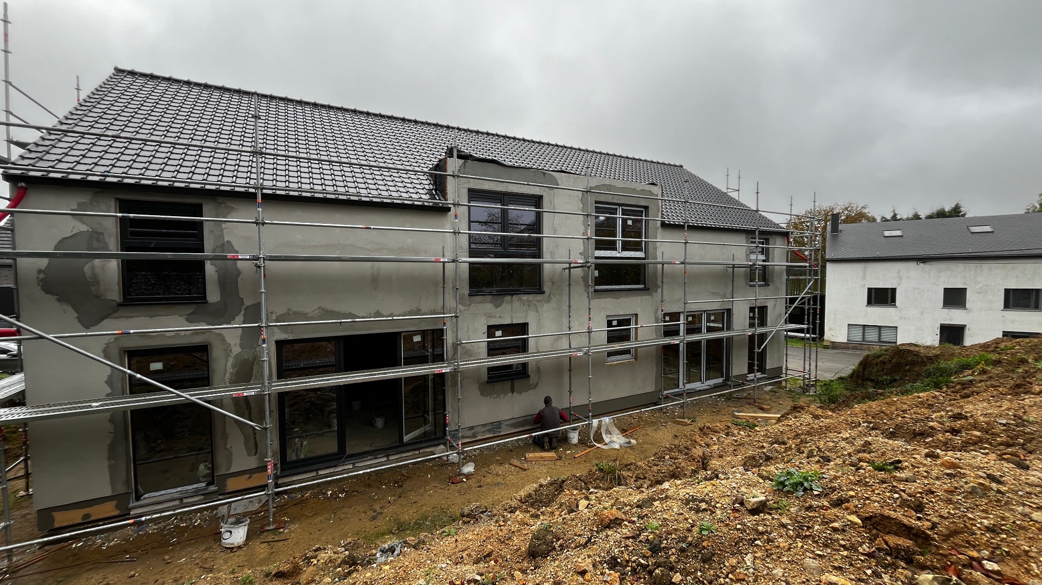 Dwupiętrowy budynek w trakcie prac wykończeniowych elewacji, z widocznymi rusztowaniami, częściowo zagruntowaną ścianą i pracownikiem przy podstawie ściany, na pierwszym planie ziemia.