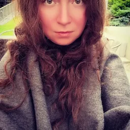 Portret kobiety o długich, kręconych brązowych włosach i jasnych oczach, ubrana w gruby szary sweter, na tle zieleni ogrodu.