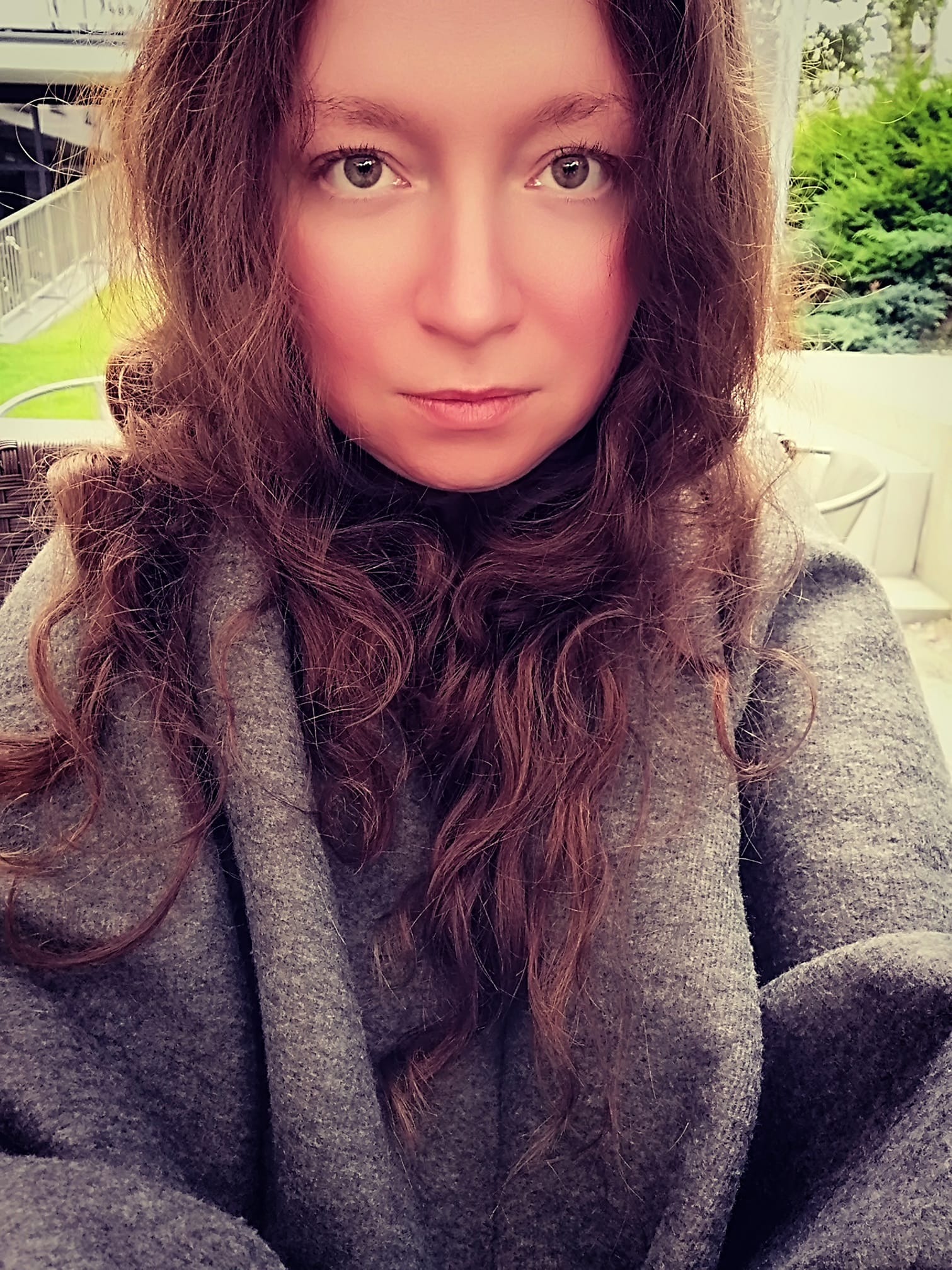 Portret kobiety o długich, kręconych brązowych włosach i jasnych oczach, ubrana w gruby szary sweter, na tle zieleni ogrodu.